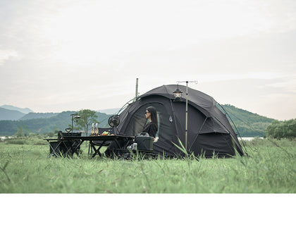 Person sitting inside a black tent in a grassy outdoor setting with mountains in the background