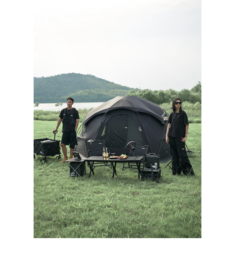 Two people standing near a camping setup with a tent, tables, and chairs in a grassy outdoor area.