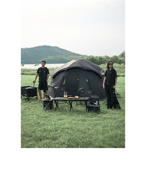 Two people standing near a camping setup with a tent, tables, and chairs in a grassy outdoor area.