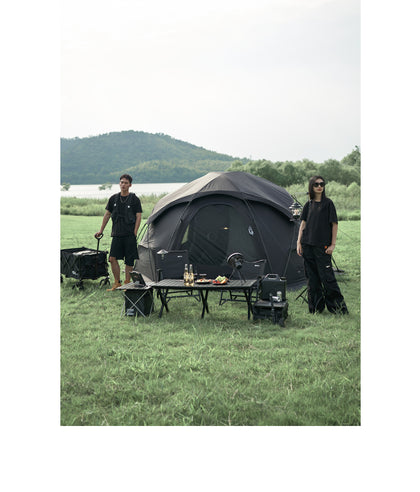 Two people standing near a camping setup with a tent, tables, and chairs in a grassy outdoor area.