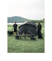 Two people standing near a camping setup with a tent, tables, and chairs in a grassy outdoor area.