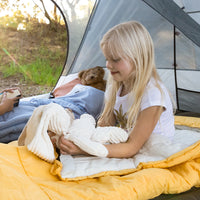 Two children in a tent, one reading a book and the other holding a teddy bear, with a natural outdoor setting.
