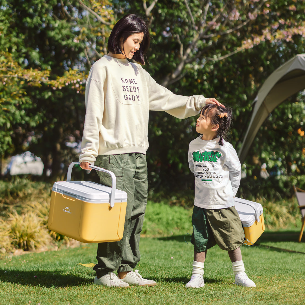 An adult and a child are outdoors with a yellow cooler bag. The adult is reaching for the child, who is wearing a grey sweater with green writing and a green pants, standing on one foot.