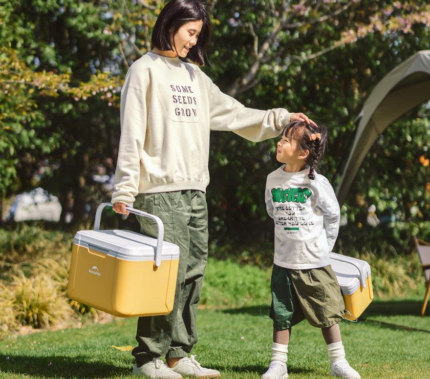 An adult and a child are outdoors with a yellow cooler bag. The adult is reaching for the child, who is wearing a grey sweater with green writing and a green pants, standing on one foot.