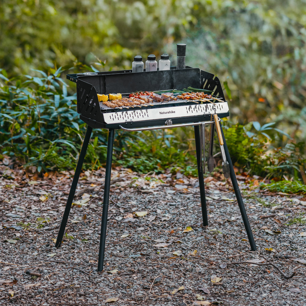 Naturehike vertical BBQ grill in black with a porcelain charcoal basin, set on a natural outdoor setting with food items on the grill and grilling tools placed on top.