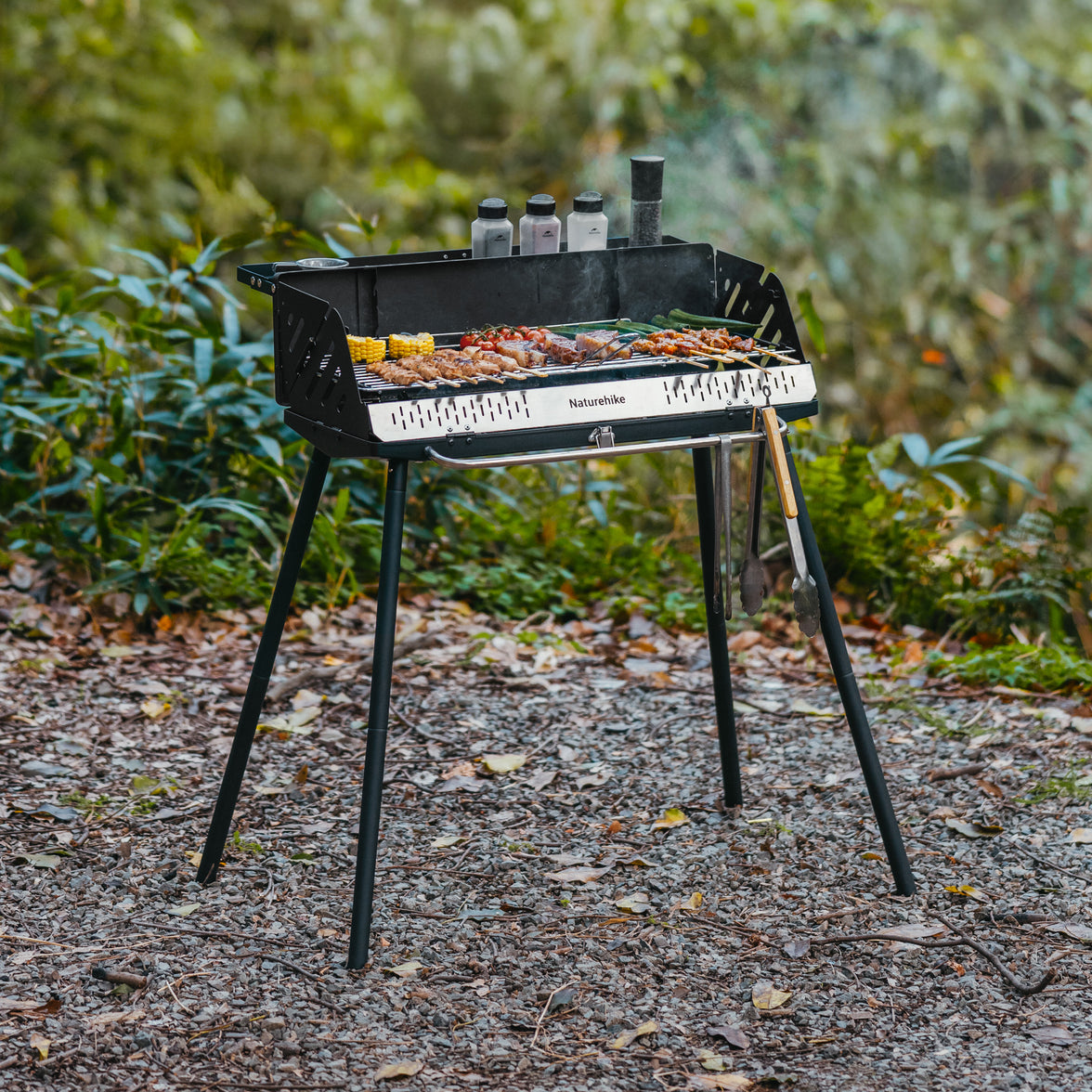 Naturehike vertical BBQ grill in black with a porcelain charcoal basin, set on a natural outdoor setting with food items on the grill and grilling tools placed on top.