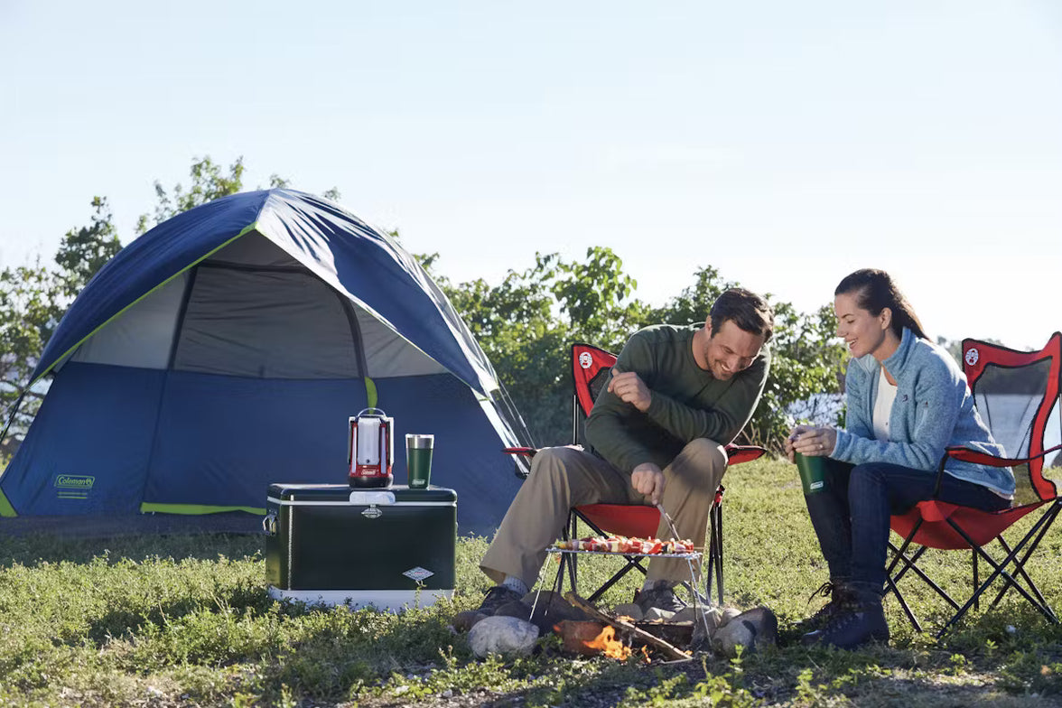 Two people camping outdoors, cooking on a grill in front of a blue tent.