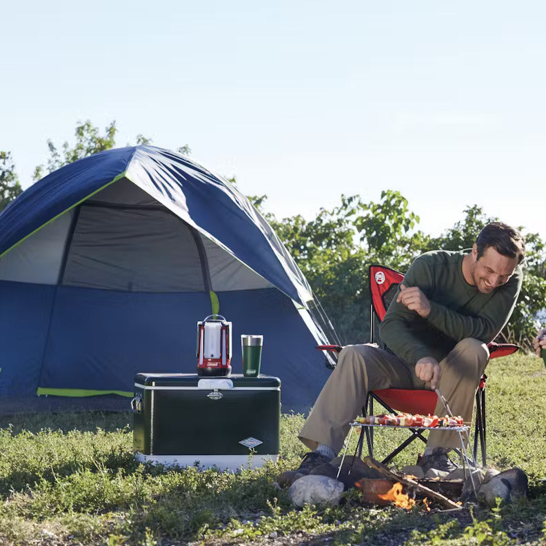 Two people camping outdoors, cooking on a grill in front of a blue tent.