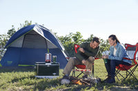 Two people camping outdoors, cooking on a grill in front of a blue tent.