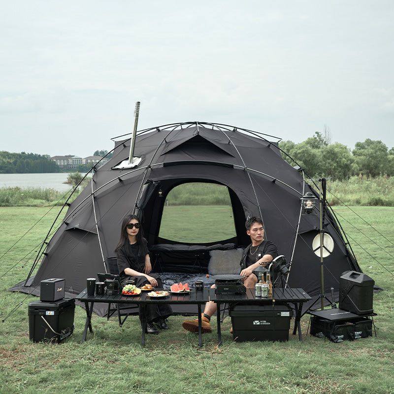 Two people sitting outside a large black tent in a grassy area with a lake in the background.