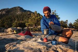 Person camping in the mountains, preparing food over a small stove.