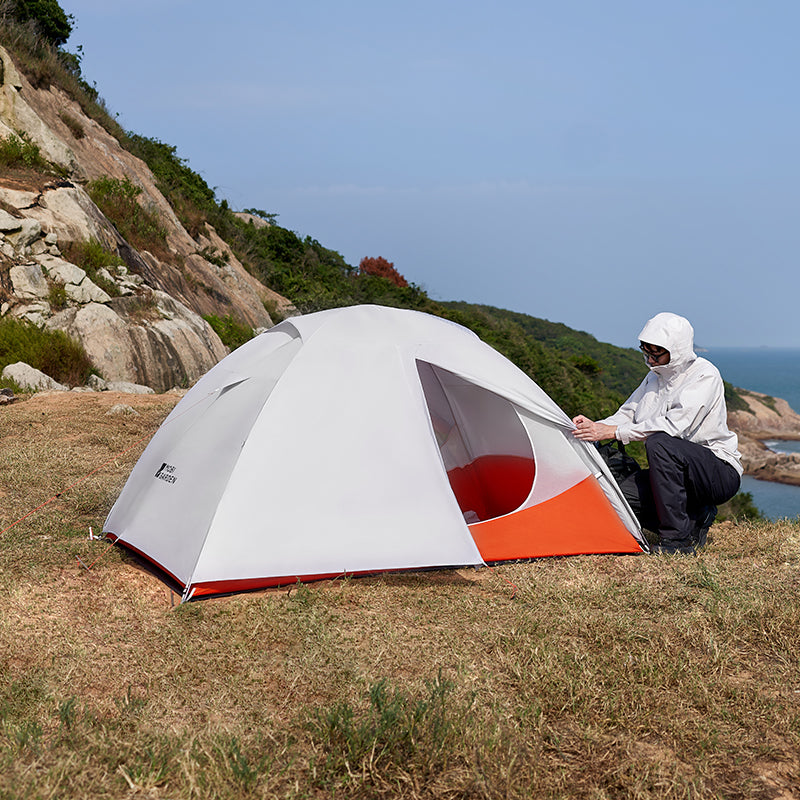 Person setting up a dome-shaped tent in a scenic outdoor location with mountains and water in the background.