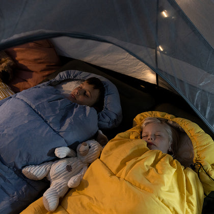 Two children sleeping in a tent with blue and yellow sleeping bags, surrounded by camping gear.