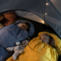 Two children sleeping in a tent with blue and yellow sleeping bags, surrounded by camping gear.