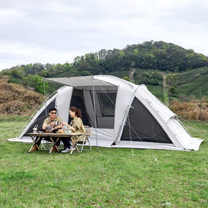 Two people sitting at a table outside a large white tent in a grassy field with trees in the background.