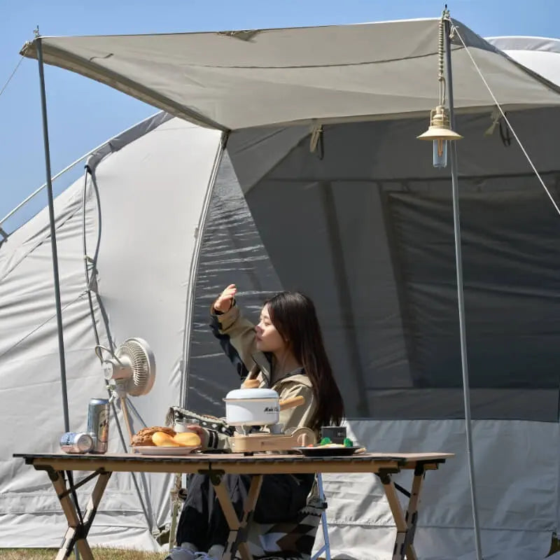Person sitting at a table in front of a tent with camping gear and food.