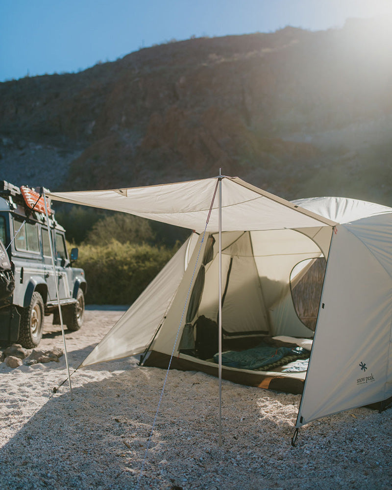 Snow Peak Alpha Breeze set up beside a Land Rover on gravel, with a tarp extended off the front.