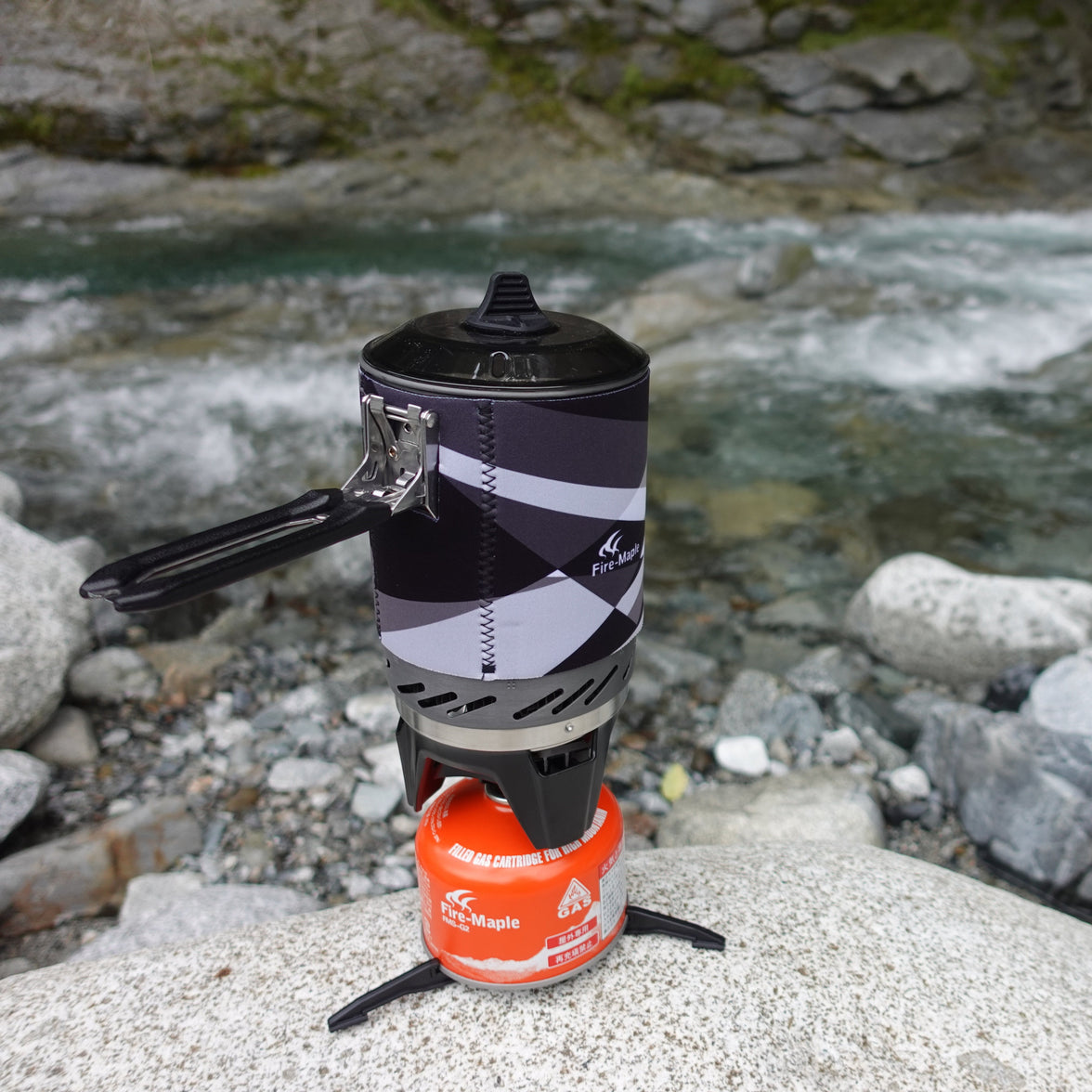 A portable cooking system, with a black and silver pot on top of a red camping stove, placed on a rock near a river.