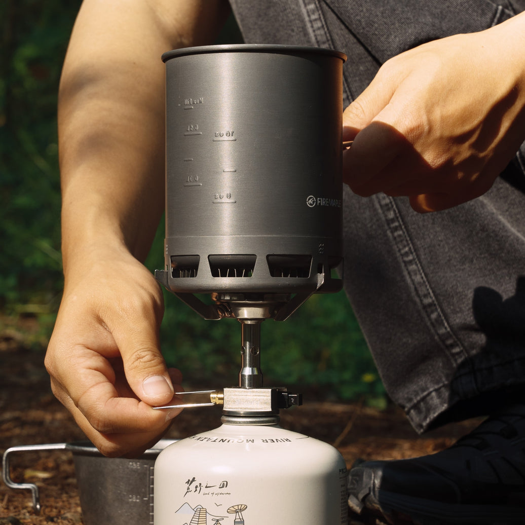 A person is adjusting a portable camping stove with a gray pot on top, which is connected to a gas canister.