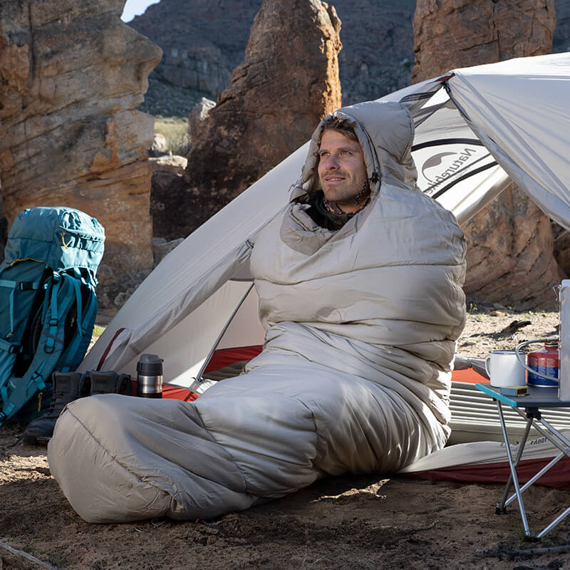Person wrapped in a sleeping bag outdoors with camping gear around
