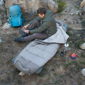 Person camping in a desert setting with a sleeping bag, backpack, and cooking equipment.