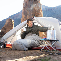 Man camping in a desert area, sitting inside a tent with a view of rocky cliffs.
