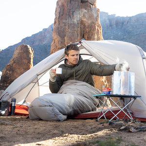 Man camping in a desert area, sitting inside a tent with a view of rocky cliffs.