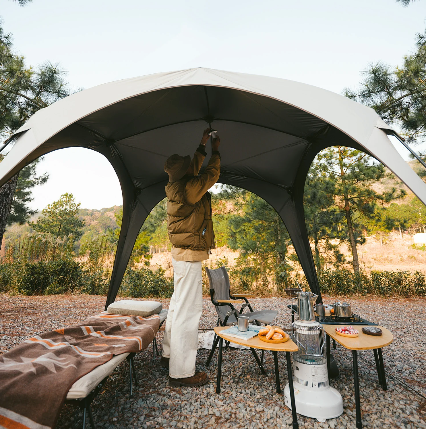 Person setting up a large outdoor canopy in a camping area with trees and nature in the background.