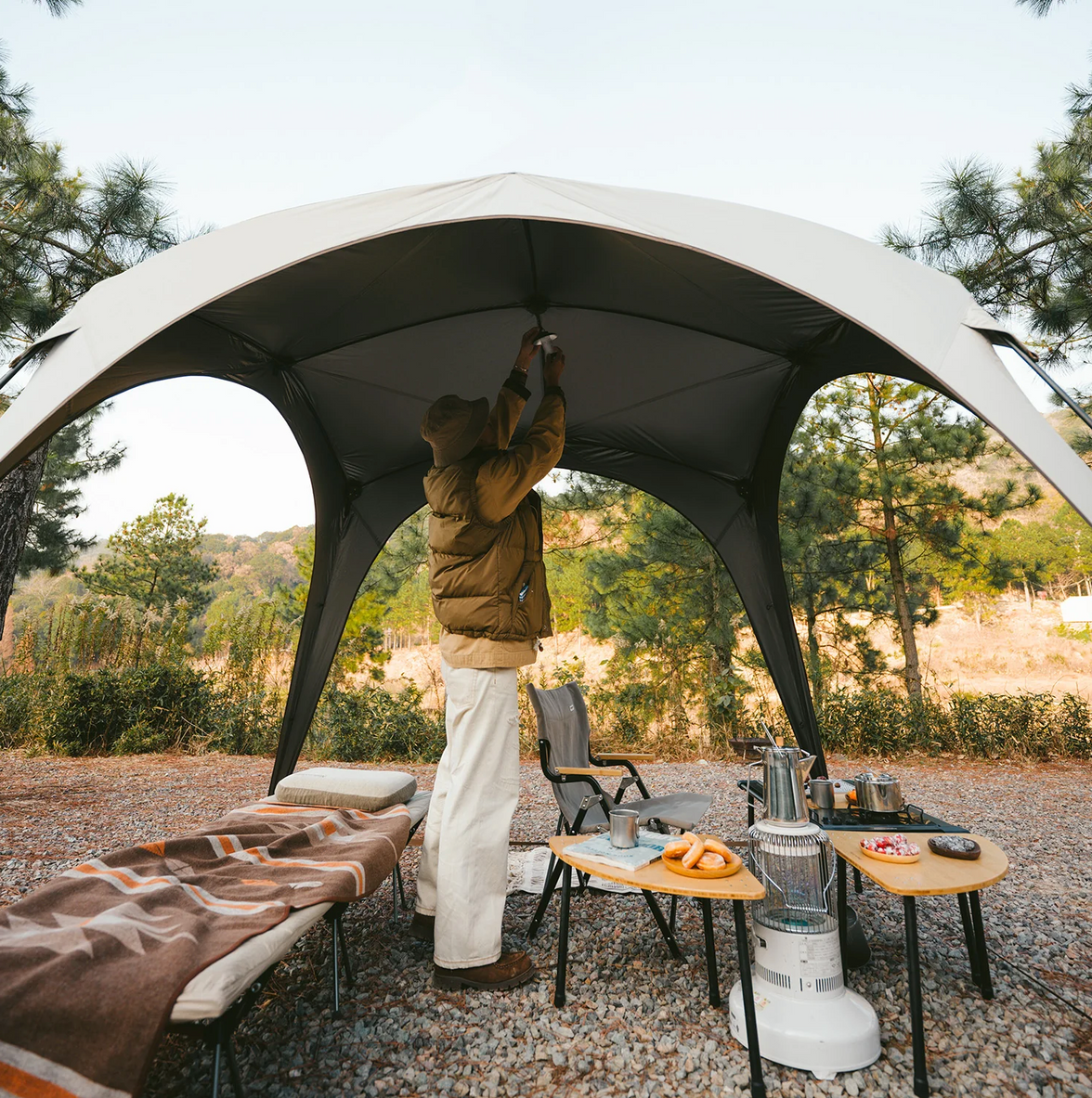 Person setting up a large outdoor canopy in a camping area with trees and nature in the background.