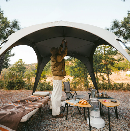 Person setting up a large outdoor canopy in a camping area with trees and nature in the background.