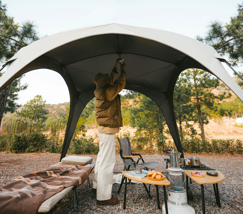 Person setting up a large outdoor canopy in a camping area with trees and nature in the background.