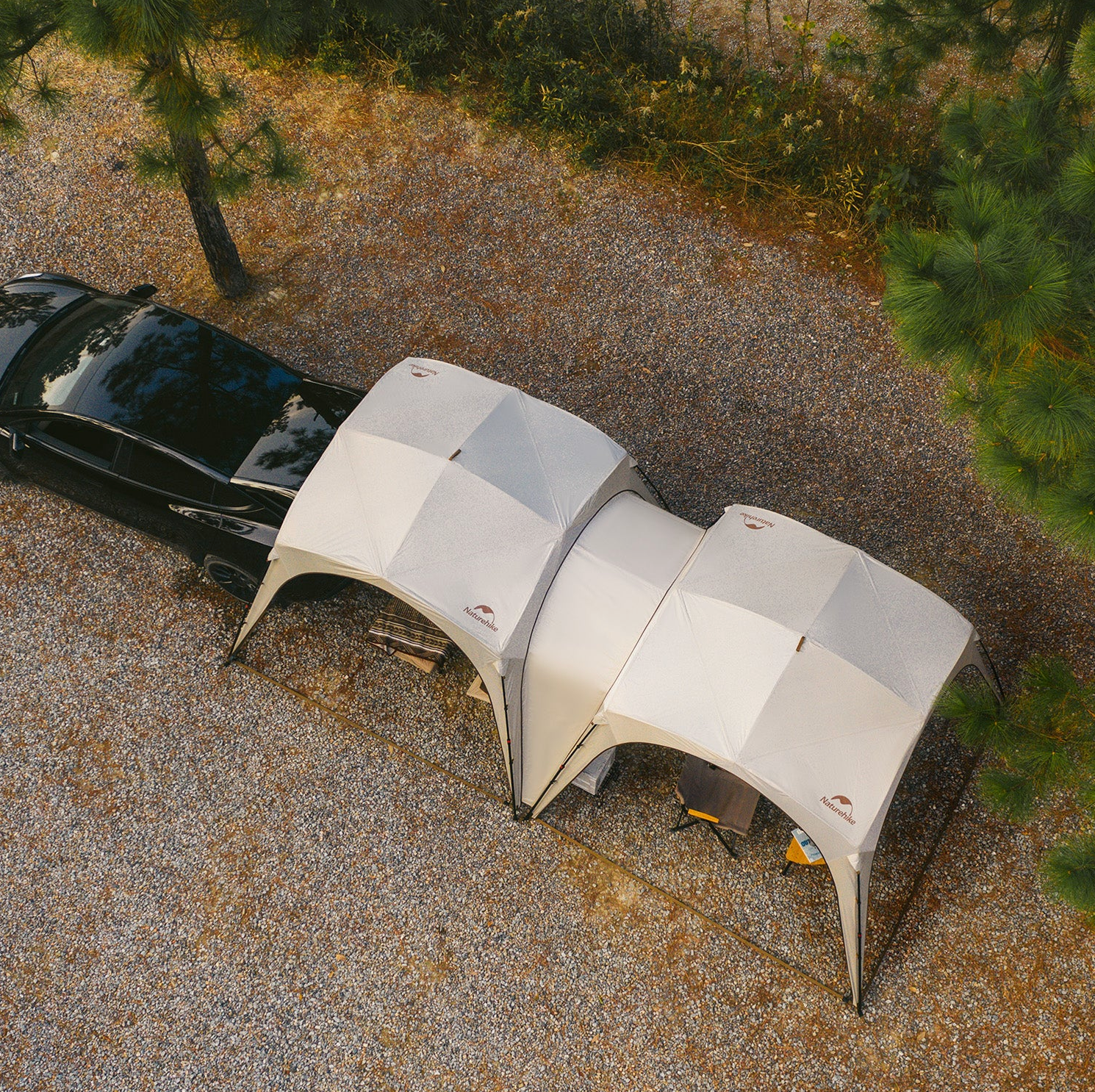 Two dome-shaped tents attached to a black car in a gravel area with trees.