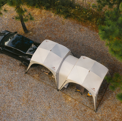 Two dome-shaped tents attached to a black car in a gravel area with trees.