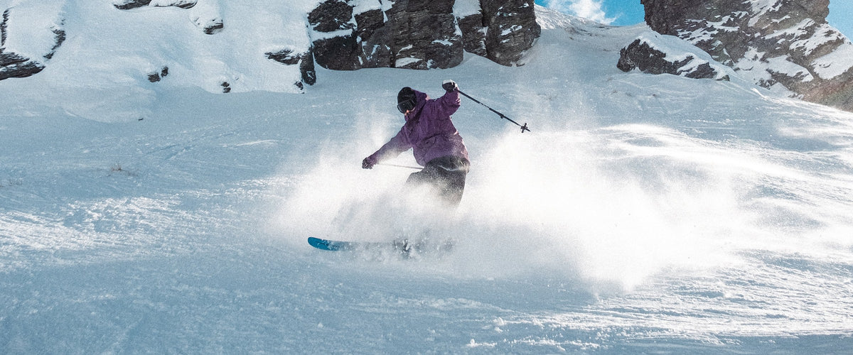 Skier in a purple Capelin Crew shell turning in powder below snowy rocks