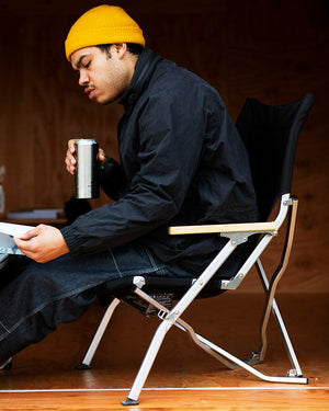 Snow Peak Shimo Can Cooler 500 held by a seated man in a yellow beanie, can inside, Snow Peak folding chair in frame