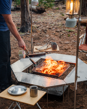 Snow Peak Folding Torch GT-120 in hand lighting split wood in a fire pit at a dusk campsite, lantern glowing