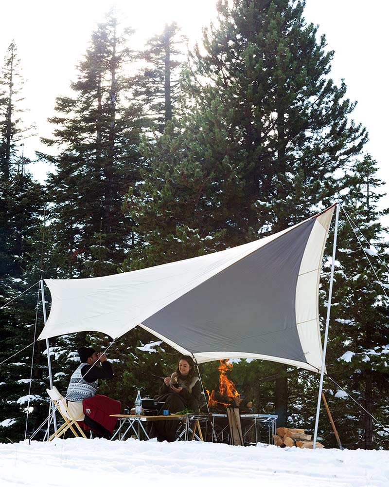 Snow Peak Takibi Tarp Hexa Set M in ivory with grey inner roof, pitched on snow in a conifer forest, two campers by a fire