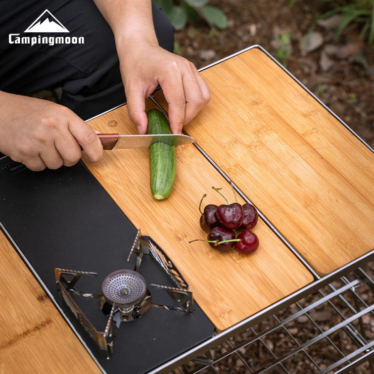 Person cutting a cucumber on a wooden cutting board with a knife, surrounded by fruits on a camping stove.