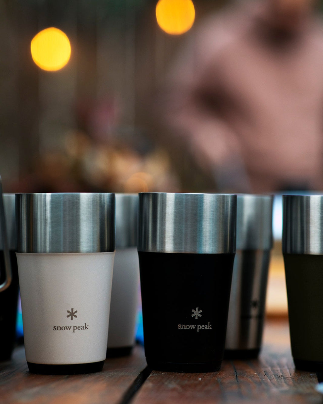 Row of Snow Peak Shimo Tumblers in ivory, black, stainless and olive on a wooden table under warm string lights