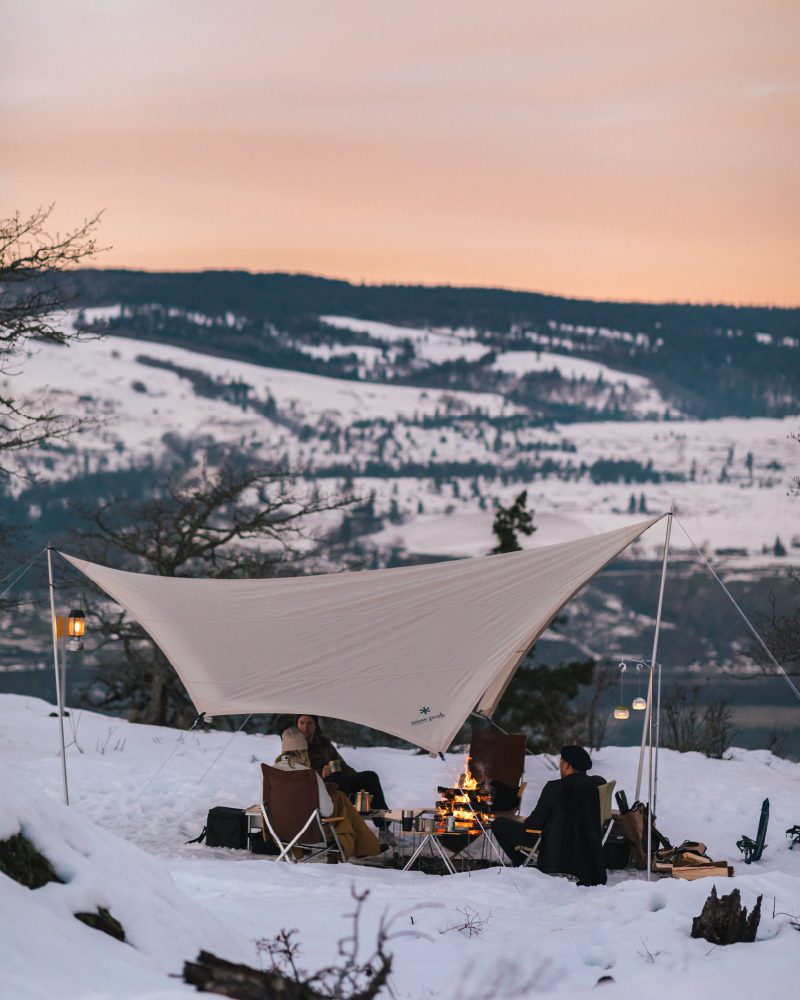 Snow Peak Takibi Tarp Hexa Set M in ivory on a snowy ridge at sunset, two campers by a fire with lanterns on stands