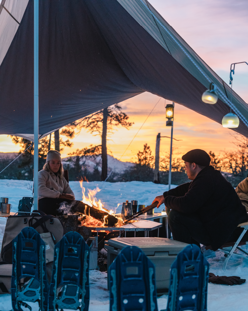 Snow Peak Takibi Tarp Hexa Set M at dusk on snow: two campers by an open fire, hanging lanterns, snowshoes in the foreground