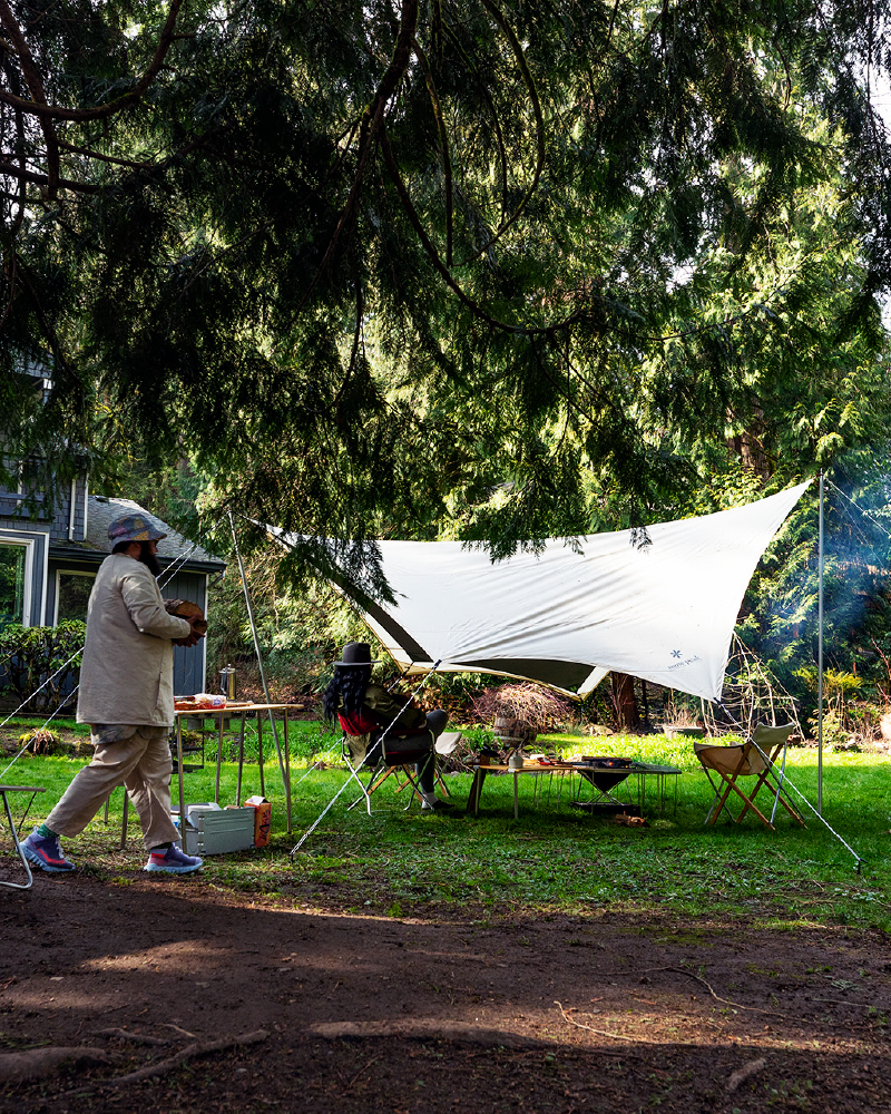 Snow Peak Takibi Tarp Hexa Set M in ivory pitched under a cedar in a backyard, campers at low tables, firewood being carried