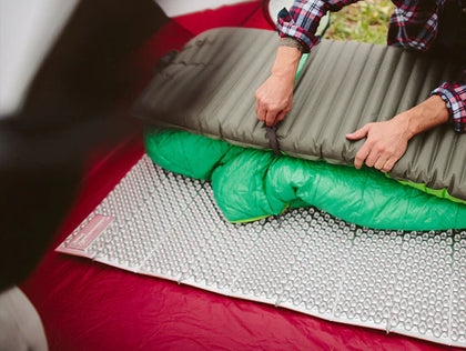 Person setting up a camping mattress on a bubble mat outdoors.